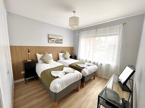 A hotel room with two beds, a bedside table, a lamp, a window with curtains, and a painting on the wall. - Luxury By The Coast 7 (Galway)