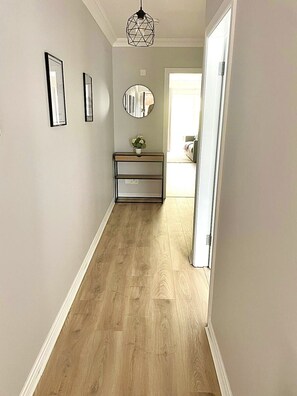 A hallway with a wooden floor, a white door, a console table with a mirror, and a hanging light fixture. - Luxury By The Coast 7 (Galway)