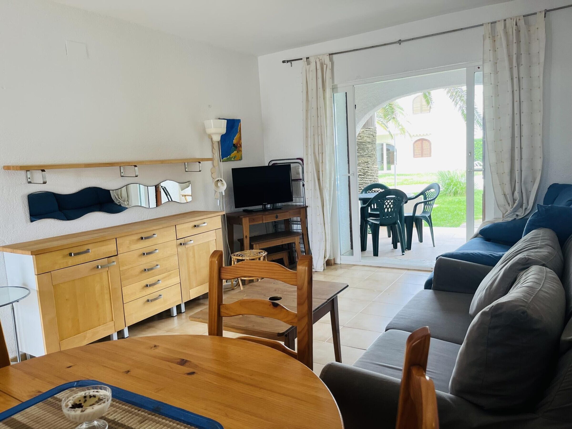 Ein Zimmer mit einem Holzesstisch, einer Couch, einem Fernseher und Blick ins Freie.