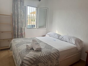 A neatly made bed with a patterned bedspread, a pair of folded towels, and a window with a view of buildings outside. - Apartment in Denia - Perfect for Your Vacation (Denia)