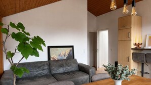 Ein modernes Wohnzimmer mit grauem Sofa, einem Holzesstisch mit gehäkelter Tischdecke, einer Topfpflanze und einem gerahmten Bild an der Wand.