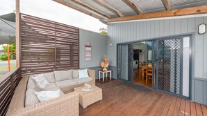 A covered outdoor seating area with a sofa and a small table, a wooden deck, and a sliding glass door leading to an interior space. - Sandy Shores, Serenity Resort (Diamond Beach)