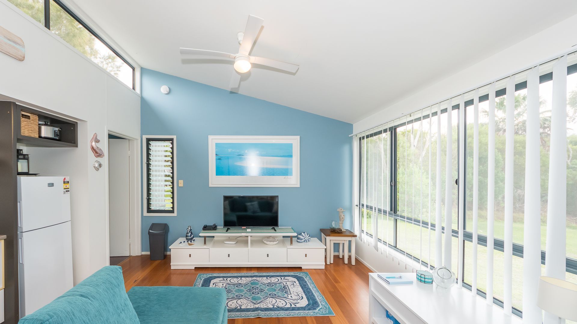 A modern living room with a flat-screen TV, a ceiling fan, and a large window with blinds.