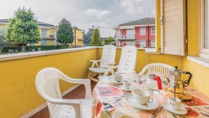 Un balcón con sillas de plástico blanco, una mesa para dos con tazas y una cafetera, y vistas a edificios amarillos y árboles.