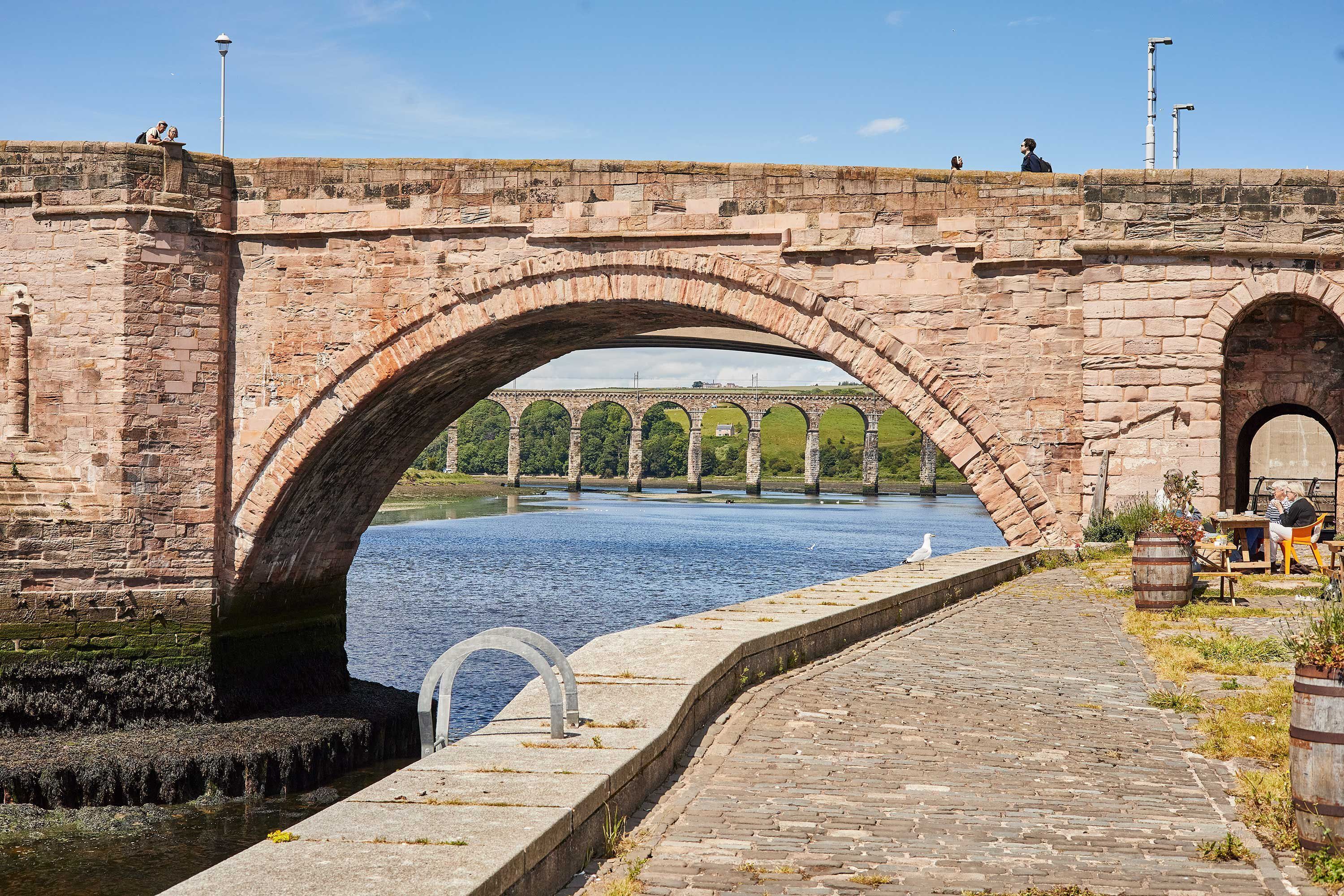 Un pont de pedra amb arcs sobre una massa d'aigua, una passera pavimentada i una vista llunyana d'un paisatge verd.