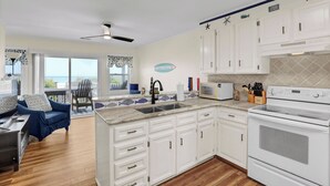 Una cocina con gabinetes blancos, una estufa y un microondas. Hay una zona de comedor con un sofá azul y una silla.