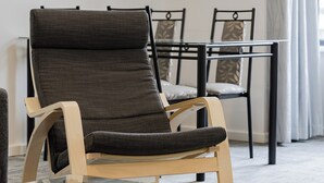 A modern living room with a dark upholstered armchair, a glass coffee table, a black and white cityscape painting, and a dining area with chairs and a table. - Your East Perth Haven awaits! Walk to Optus Stadium, Crown Perth and the CBD. Enjoy iconic Swan River walks, vibrant Claisebrook Cove for coffee and dining and super easy city access with the free CAT bus (20mins). (East Perth)