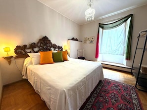 Ein Schlafzimmer mit einem großen Bett, einer Chandelieber, einem fensterbepften Fenster, Holzboden und einer Stehlampe.