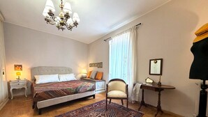 Quarto com duas camas, um candelabro, uma cadeira, uma escrivaninha e uma janela grande com cortinas.