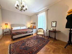 Ein Schlafzimmer mit zwei Betten, einer Kandelaber, einem Sessel, einem Schreibtisch und einem großen Fenster mit Vorhängen.
