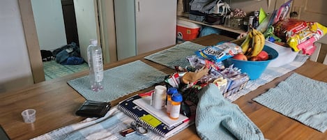 Una habitación desordenada con una mesa de madera, varios objetos encima, un espejo y una estantería con utensilios de cocina.