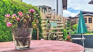 Een rustieke buitenomgeving met een houten tafel, een glazen vaas met roze bloemen, een houten hek en een gebouw met een bord in de achtergrond.