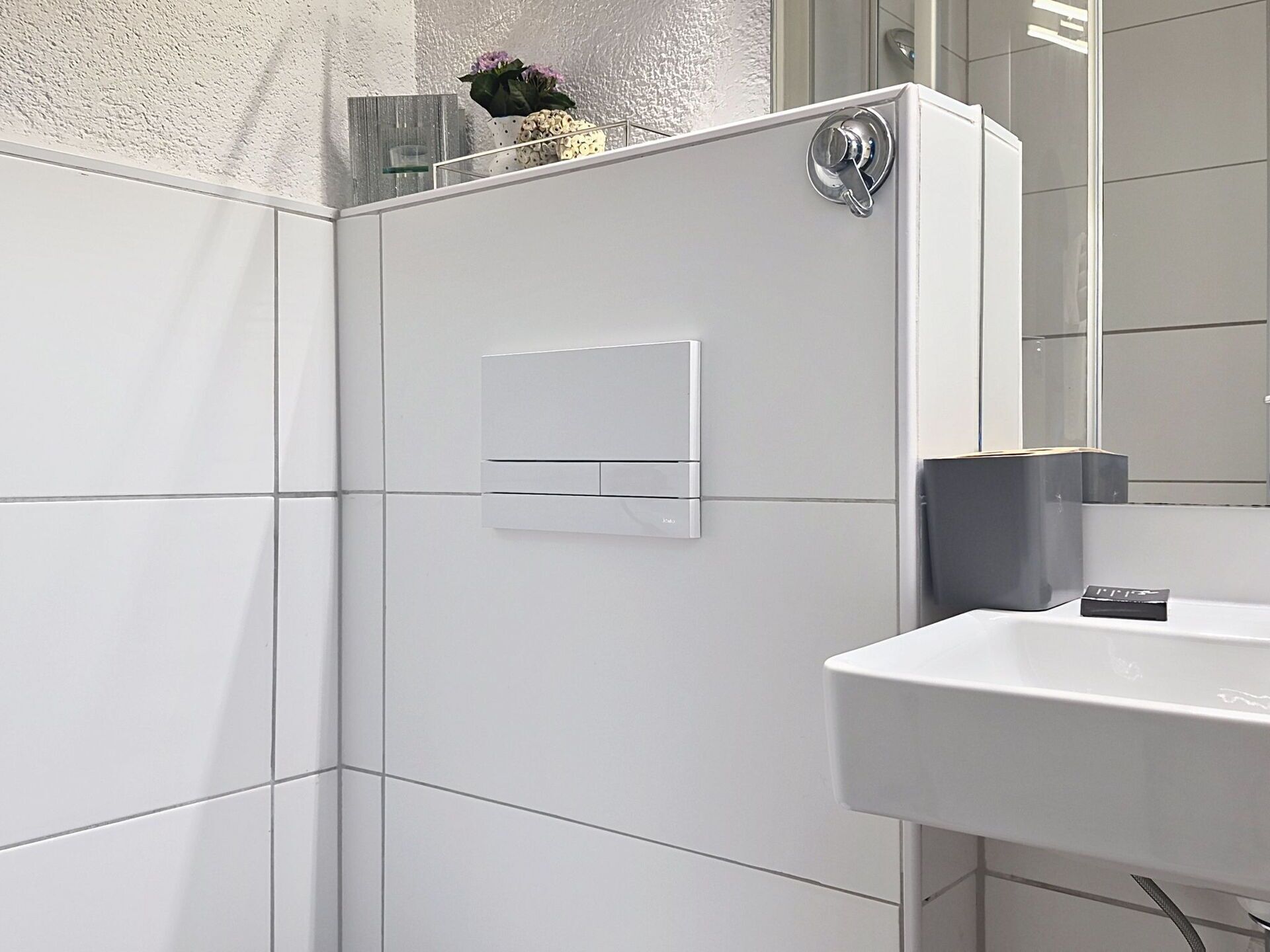 A bathroom with white tiles, a white sink, a mirror, and a shower control panel.