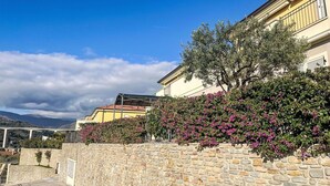 Un muro de piedra con jardín, un edificio amarillo con balcón y cielo despejado.