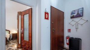 Un pasillo con suelo de mosaico, una puerta de madera, una cama en la habitación contigua y un extintor en la pared.