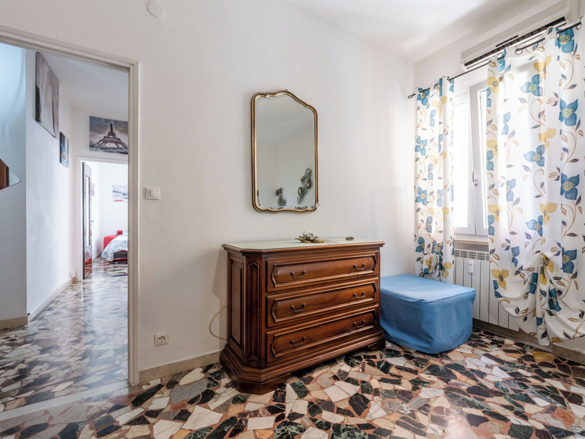 Une pièce avec une commode en bois, un miroir, un banc bleu, un rideau à motifs et un sol carrelé.