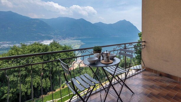 Een balkon met een tafel, stoelen en uitzicht op een meer en bergen.