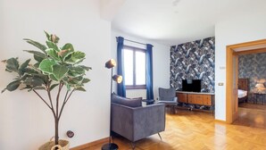 Een moderne woonkamer met een grote plant, een grijze bank, een bureau met een computer en behang met een patroon.