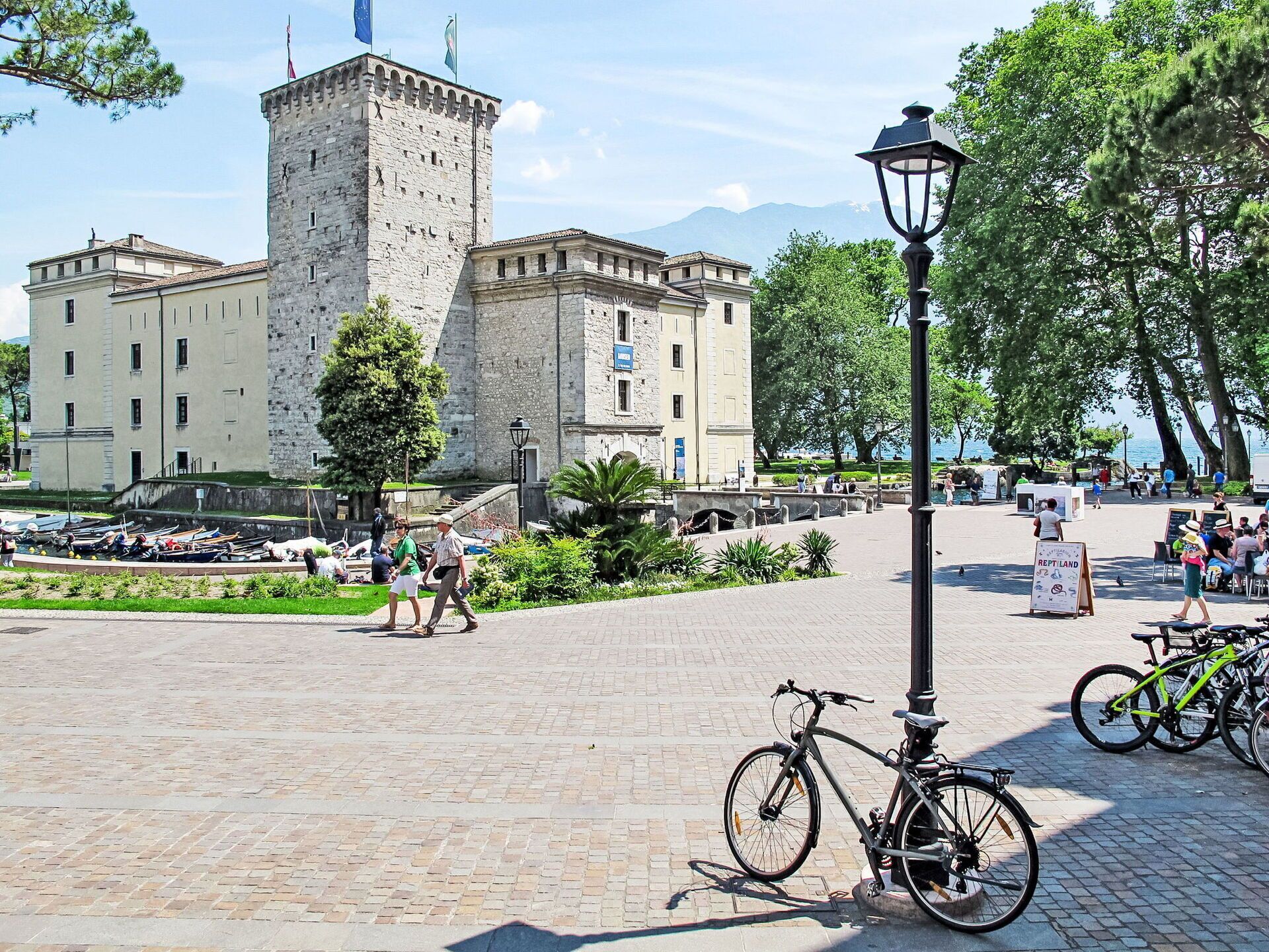 Un bâtiment historique en pierre, doté d’une tour, une place pavée où l’on aperçoit des vélos, et un panneau indicateur.