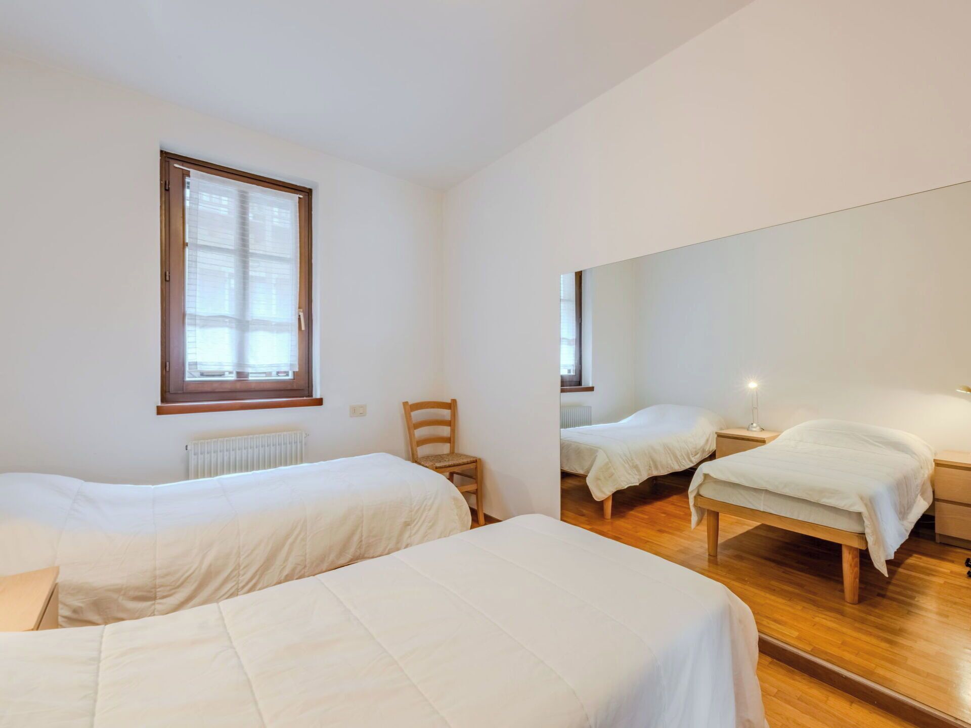 A room with two single beds, a wooden chair, a bedside table with a lamp, and a large mirror on the wall.