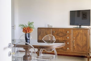 Ein Essbereich mit einem transparenten Glastisch, einer Holzkombination mit Schubladen und einem Fernseher an der Wand.