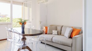 A modern living room with a round glass table, clear chairs, a beige sofa with orange cushions, and a vase with flowers on a windowsill. - Lovely apartment for 4 people with WIFI, A/C, TV, balcony and panoramic view (Lido Di Fermo)