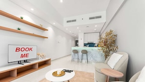 A modern living room with a TV, sofa, coffee table, and a kitchen area in the background. - Free Housekeeping Visits During Your Stay - Stayshort - Studio in Dubai Land That Sleeps 3 Close to Global Village (Dubai)