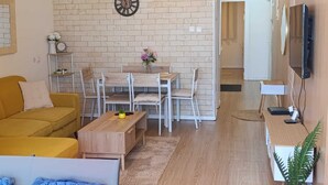 A living room with a sofa, a coffee table, a dining table with chairs, a clock on the wall, and a television on the wall. - Luxury Stunning 2BR Marina Apt with Sea View (Ashkelon)