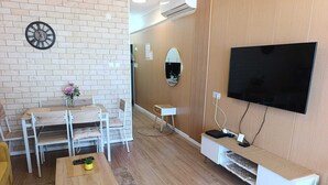 A modern living room with a brick wall, a dining area with a wooden table and chairs, a flat-screen TV mounted on a wooden panel wall, and a small white side table. - Luxury Stunning 2BR Marina Apt with Sea View (Ashkelon)