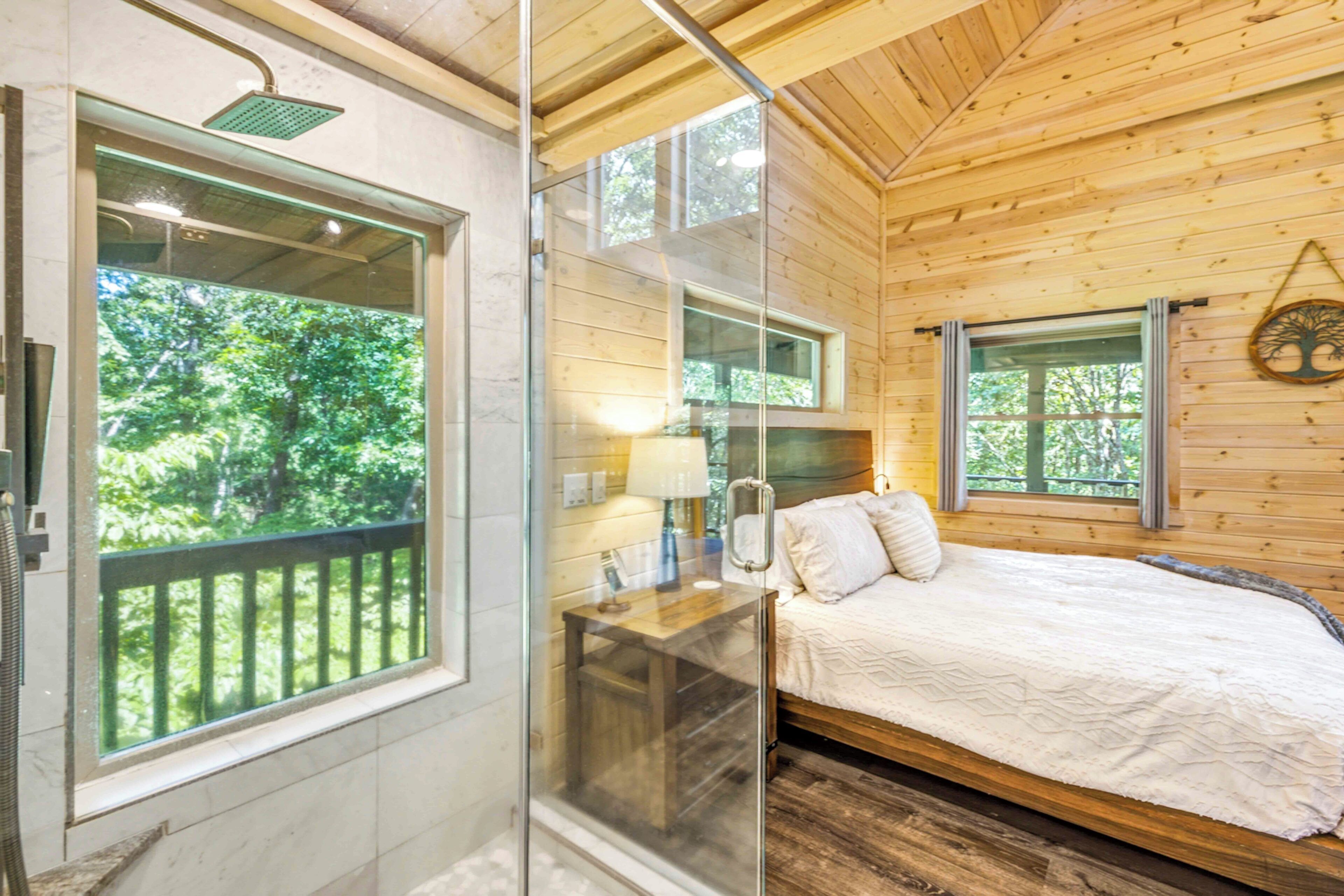 Ein Schlafzimmer einer Holzhütte mit einem Bett, einem Fenster mit Blick auf Bäume, einer Duschkabine aus Glas und einer Holzbalkendecke.