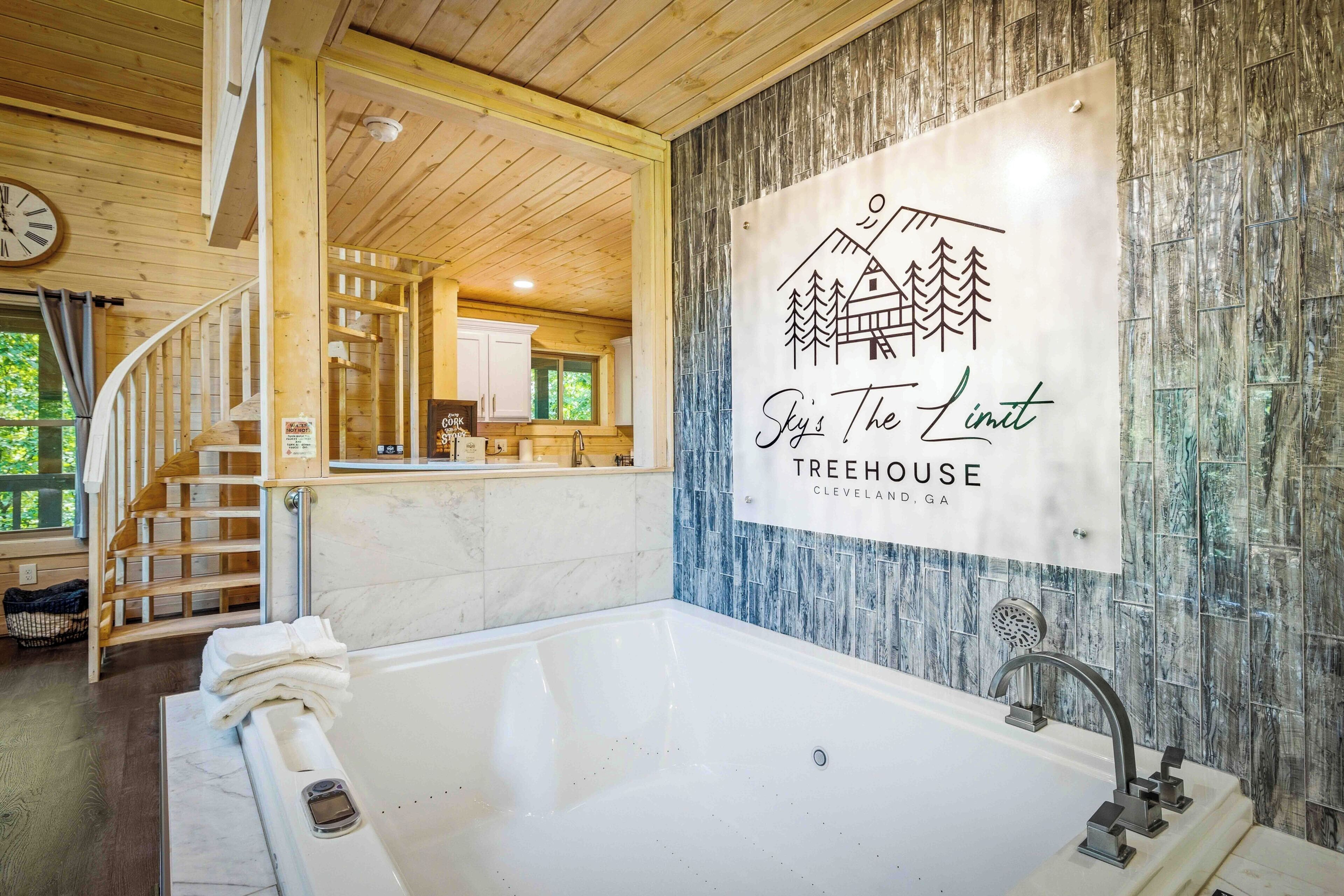 Et rummeligt badeværelse med et stort badekar, en trætrappe og et skilt med teksten 'Sky's The Limit Treehouse Cleveland, GA'.