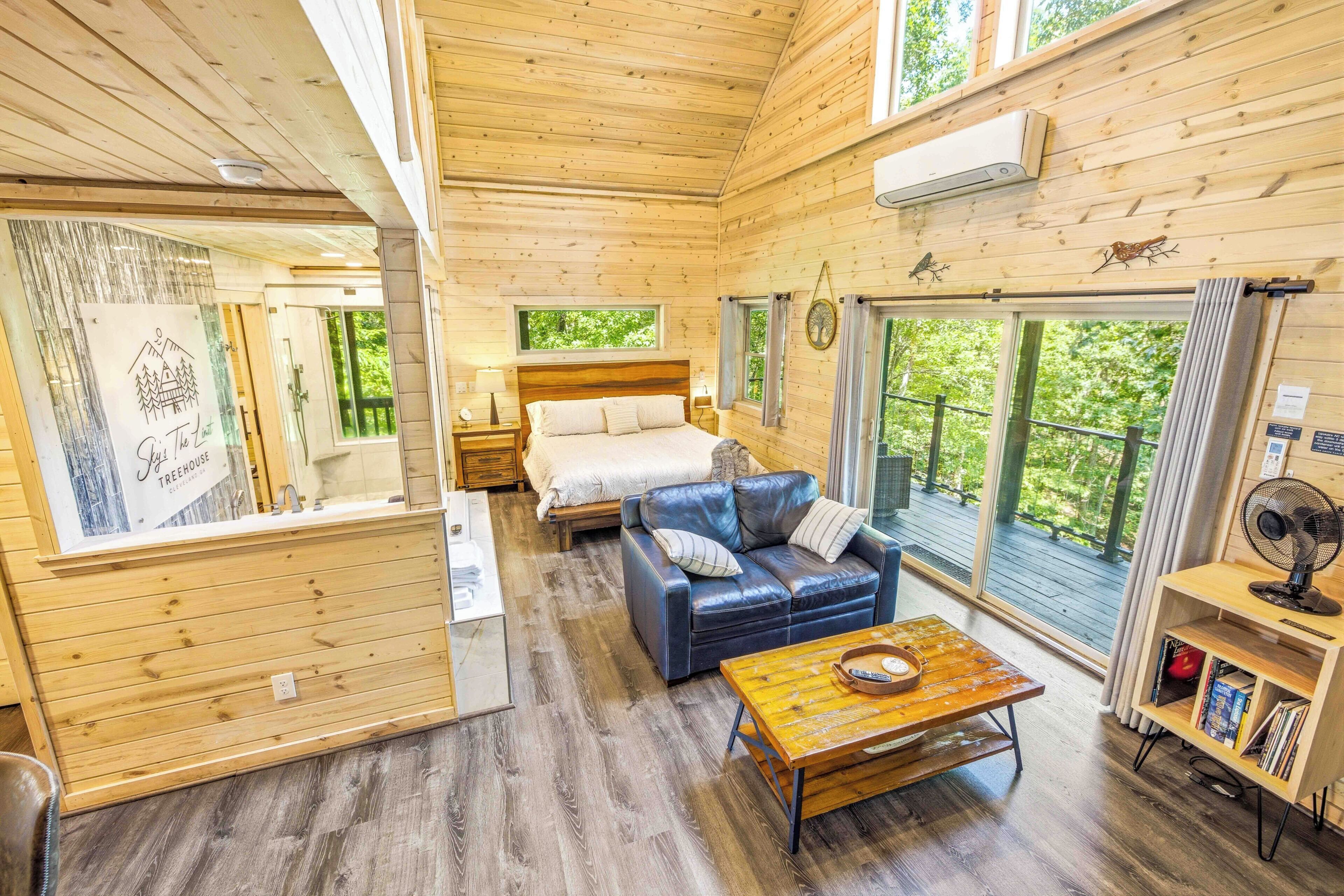Interior acollidor d'una cabana amb terra de fusta, un llit, un sofà, una taula i una finestra amb vista a vegetació.