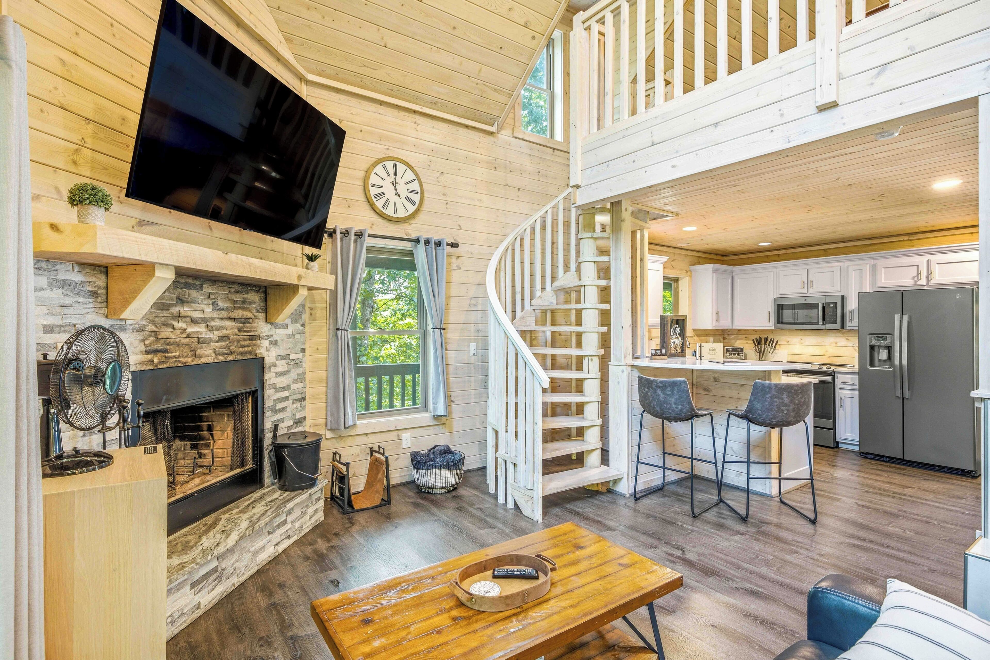 A spacious living area with a fireplace, a television mounted on the wall, a staircase leading to an upper level, and a kitchen with modern appliances.