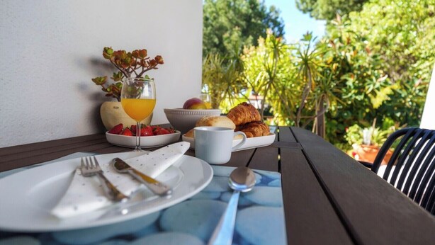 Ein Frühstückstisch mit Croissants, Erdbeeren, einem Glas Orangensaft und einer Tasse, auf einer Terrasse mit Blick auf viel Grün.