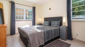 Ein Schlafzimmer mit Bett, Nachttischen, Vorhängen vor einem Fenster und Blick auf Bäume.