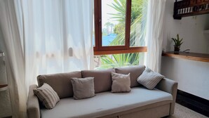 Ein beiges Sofa mit Kissen, ein Fenster mit Vorhängen, ein Holzboden und eine kleine Pflanze auf einem Regal.
