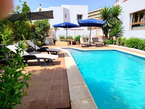 A swimming pool with lounge chairs and umbrellas, surrounded by greenery and a white building. - Villa los Robles (Eivissa)