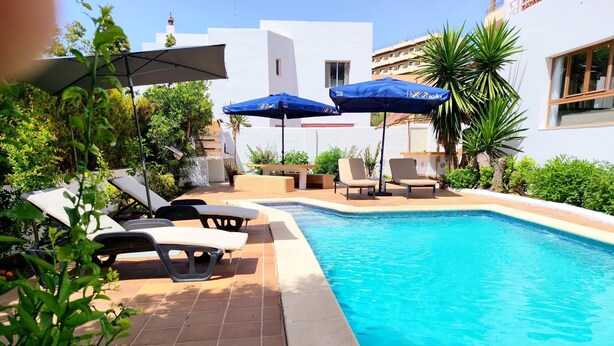 Ein Pool mit Liegestühlen und Sonnenschirmen, umgeben von Grünflächen und einem weißen Gebäude.