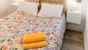 Un dormitorio con edredón estampado con motivos florales, dos almohadas blancas, cabecera tapizada en tono beige y lámparas de noche con espejo.
