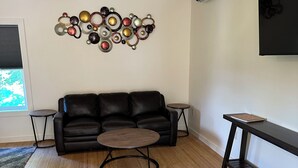 Ein Wohnzimmer mit einem schwarzen Ledersofa, einem runden Couchtisch, einer wandmontierten Klimaanlage und einem dekorativen Metallkunstwerk.
