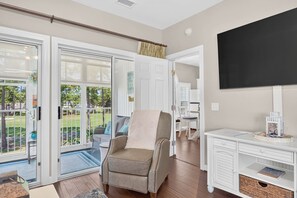 A room with a sliding glass door leading to a balcony, a sofa, a chair, a desk with a television, and a bookshelf. - Easy Breezy on the 16th Green in Sunset Beach (Sunset Beach)