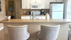 Ett kök med vita skåp, en mikrovågsugn, en spis och ett kylskåp. Det finns två barstolar med en beige klädd sits och ett dekorativt ryggstöd. En skylt med ordet ”SEA” är monterad ovanför skåpen.