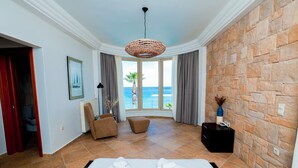 Ein Hotelzimmer mit Bett, Sessel, kleinem Tisch, Fenster mit Meerblick und Steinwand.
