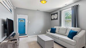 A living room with a TV, a sofa, an ottoman, a blue door, and a window with curtains.