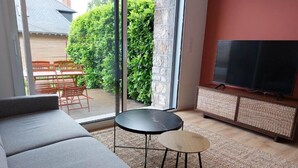 Una sala de estar con un sofá, una mesita redonda, un televisor sobre un mueble bajo y vista a un jardín a través de un ventanal.