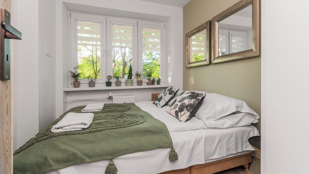 Un dormitorio con cama, ventana con persianas, espejo y estante con plantas en macetas.