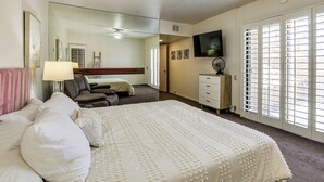 Quarto de hotel com cama, escrivaninha, cadeira, televisão e uma janela grande com persianas.