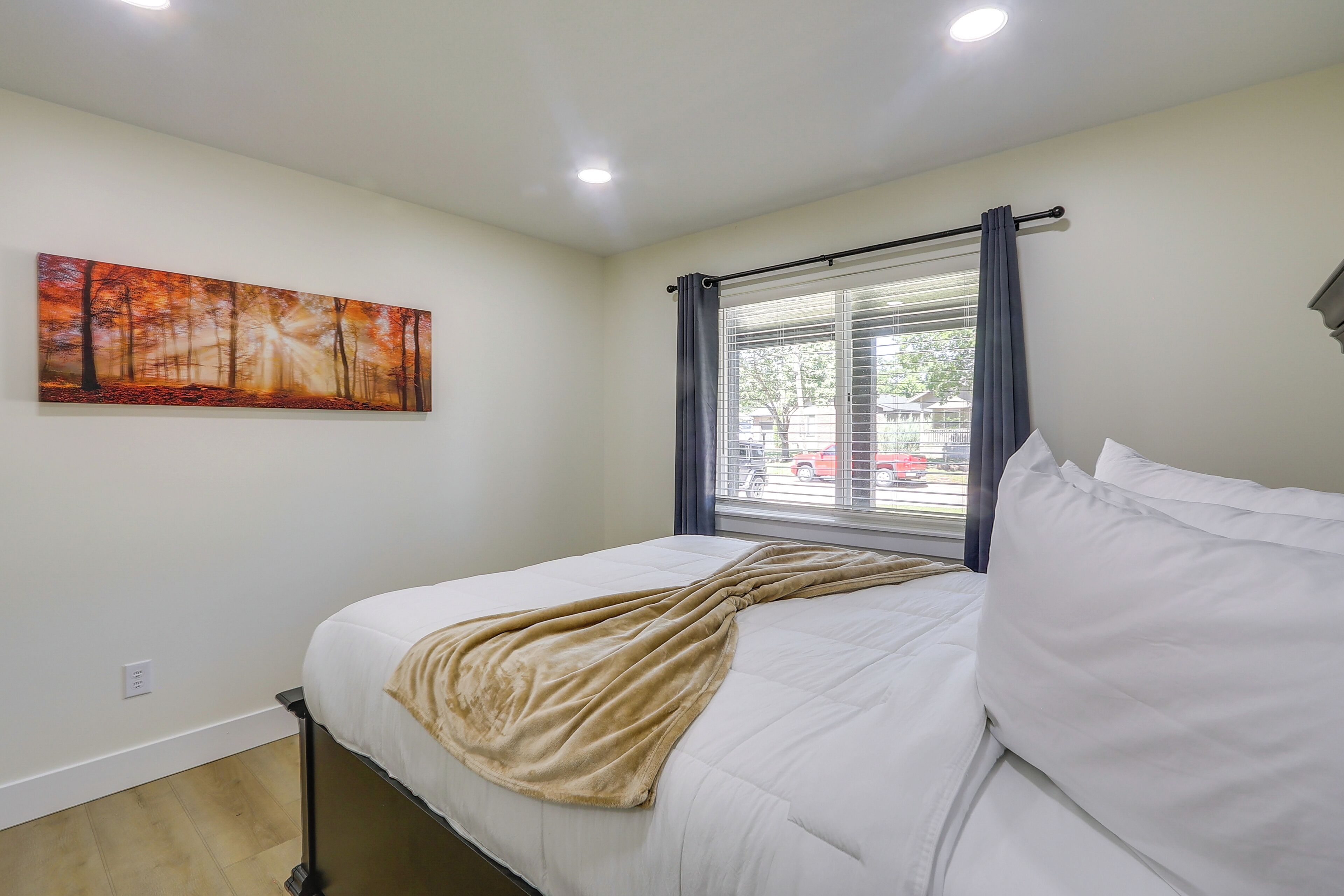 A bedroom with a bed, a window with curtains, a painting on the wall, and a ceiling light.