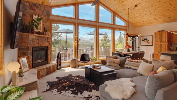 A spacious living room with a fireplace, large windows, and a ceiling fan. - Rocky Mtn Hideaway w/ Hot Tub & Panoramic Views (Fairplay)