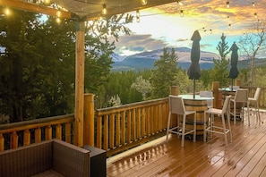 Una terraza de madera con vistas a montañas, mobiliario exterior con mesas y sillas, y luces colgantes.
